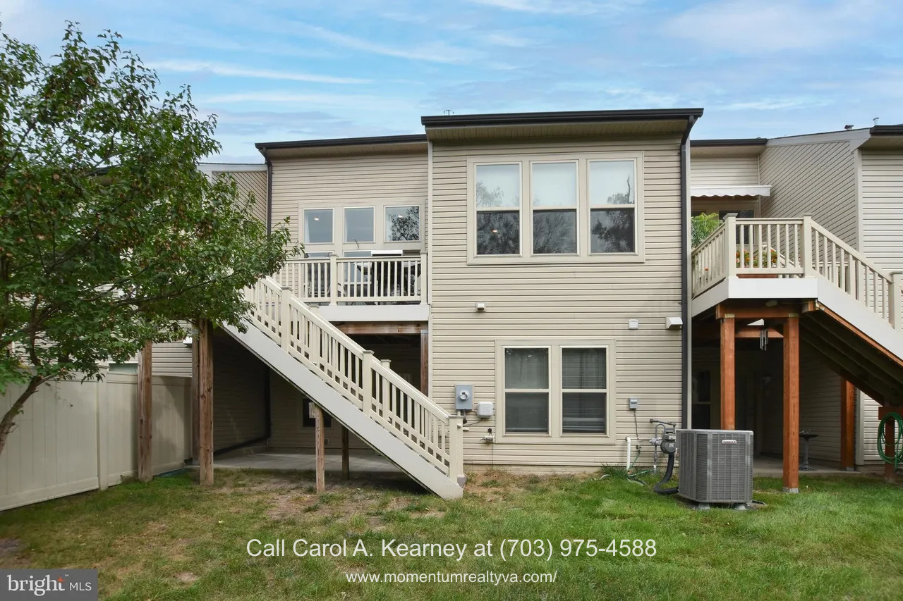 19873 Silvery Blue Ter, Ashburn, VA 20147 Rear exterior view of the townhouse at 19873 Silvery Blue Ter in Ashburn, VA, showing a main-level deck with stairs, walk-out lower level, and windows overlooking a private wooded setting in the Lexington 7 55+ community.