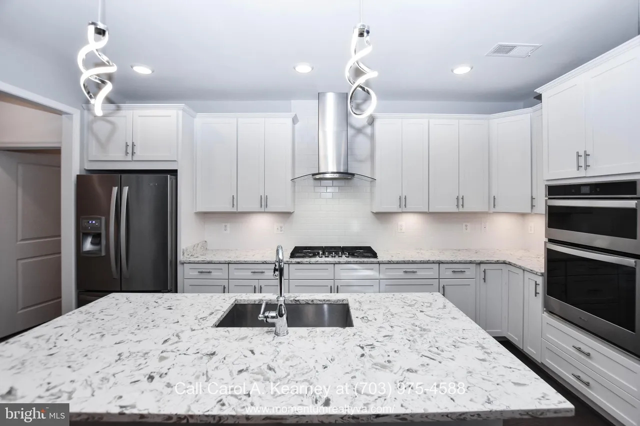 19873 Silvery Blue Ter, Ashburn, VA 20147 Gourmet kitchen at 19873 Silvery Blue Ter in Ashburn, VA, featuring white cabinetry, quartz countertops, a large center island with sink, stainless steel appliances, double wall ovens, and modern pendant lighting in the Lexington 7 55+ community.