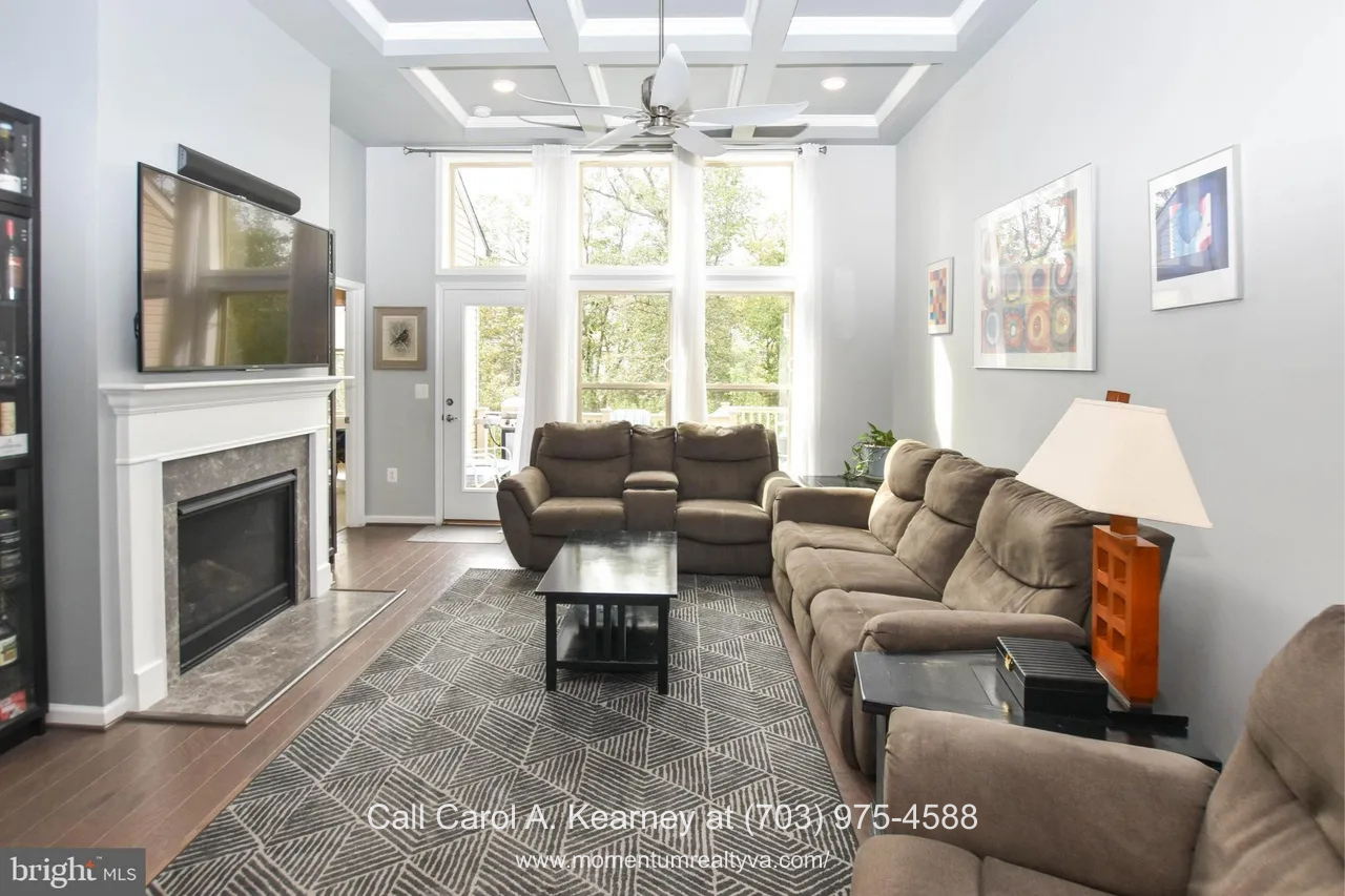 19873 Silvery Blue Ter, Ashburn, VA 20147 Bright open-concept living room at 19873 Silvery Blue Ter in Ashburn, VA, featuring a coffered ceiling with ceiling fan, large windows overlooking wooded views, gas fireplace, hardwood floors, and an inviting seating area in the Lexington 7 55+ community.