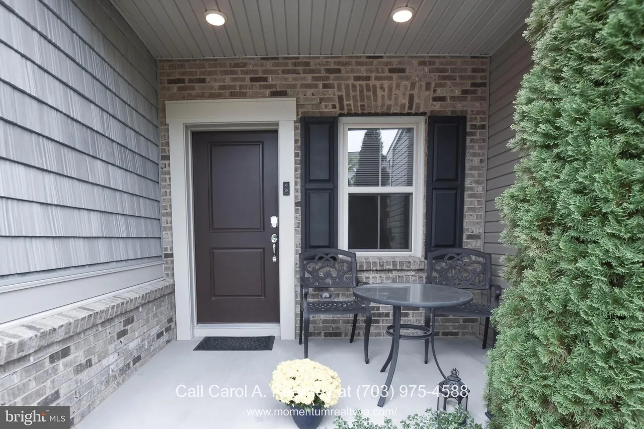 19873 Silvery Blue Ter, Ashburn, VA 20147 Covered front porch of a Lexington 7 townhouse at 19873 Silvery Blue Ter in Ashburn, VA, featuring a brick façade, dark front door, recessed lighting, and a small patio seating area overlooking landscaped greenery in a 55+ community.