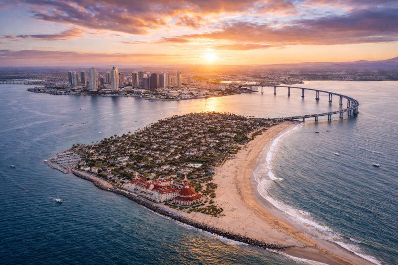 Aerial sunset view of Coronado Island showing the sandy ocean beach, the Coronado Bridge, and the downtown San Diego skyline across the bay.